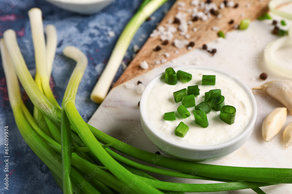 Board with bowl of tasty sour cream and sliced green onion on blue background, closeup