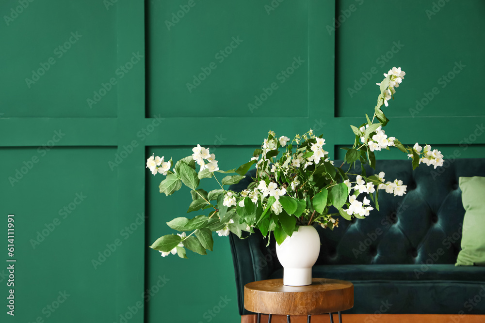 Vase with blooming jasmine flowers on coffee table and sofa near green wall