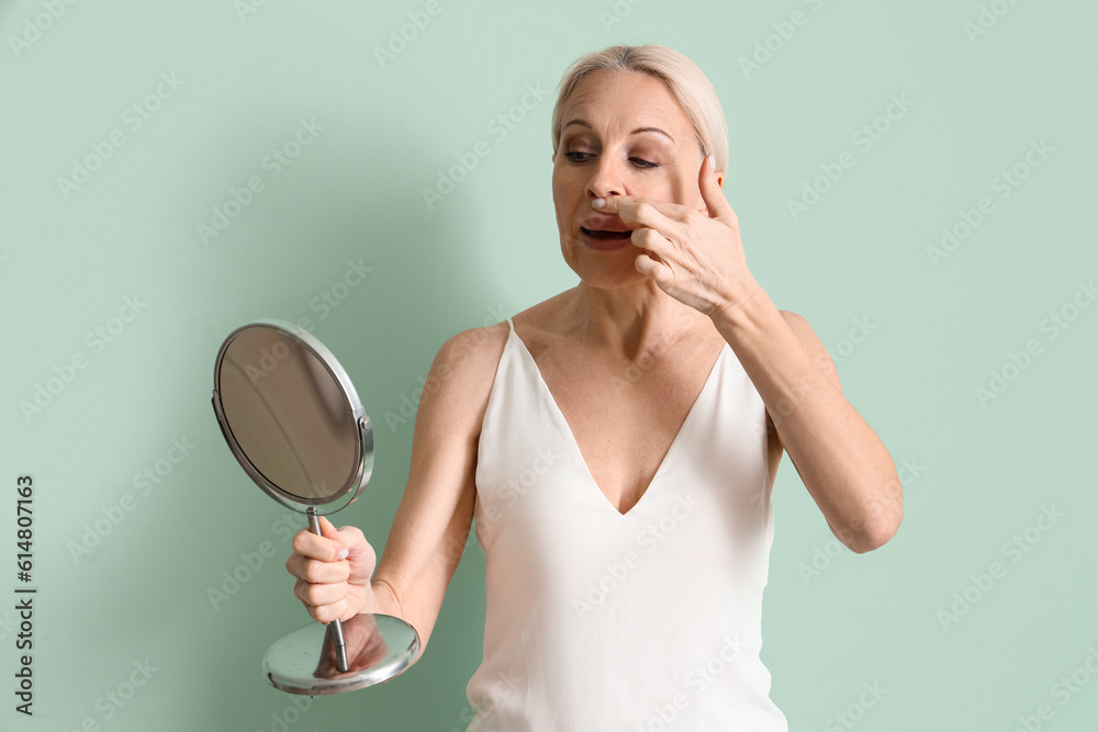 Mature woman with mirror doing face building exercise on green background