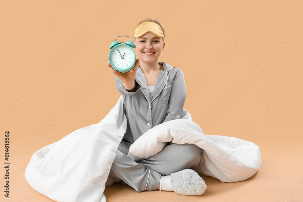 Young woman with alarm clock and blanket on beige background