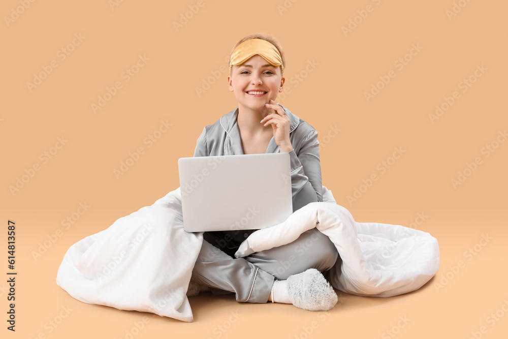 Young woman with sleeping mask and blanket using laptop on beige background