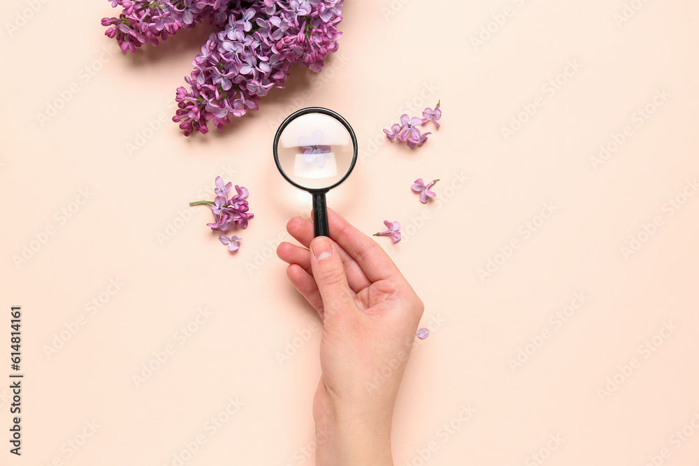 Female hand with magnifier and beautiful lilac flowers on pale pink background