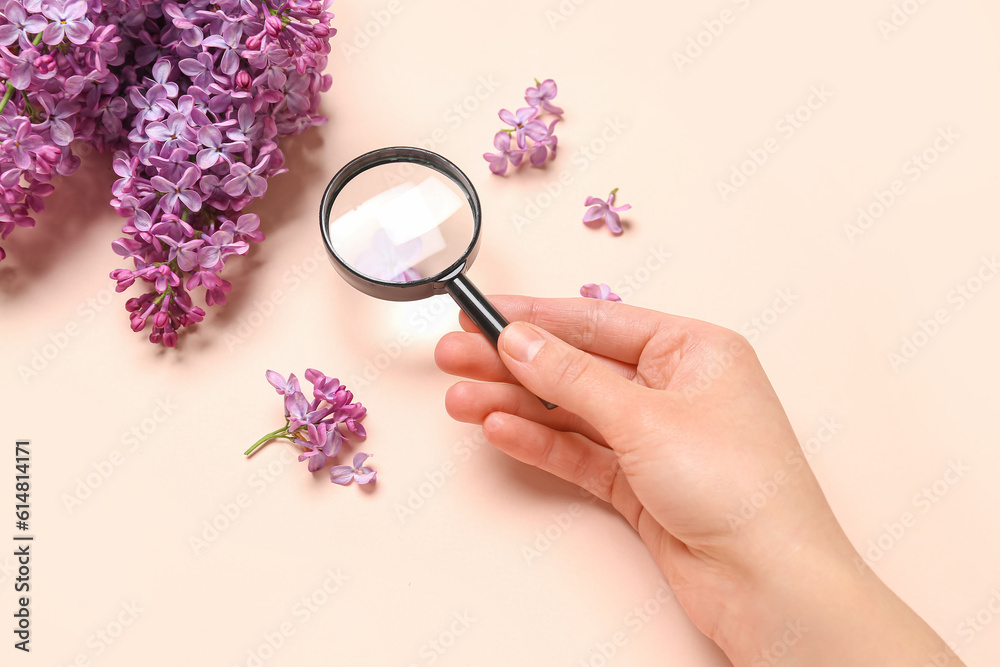 Female hand with magnifier and beautiful lilac flowers on pale pink background