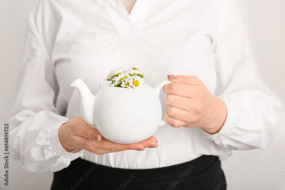 Beautiful young woman with teapot of chamomile tea and flowers near white wall