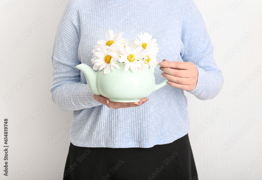 Beautiful young woman with teapot of chamomile tea and flowers near white wall