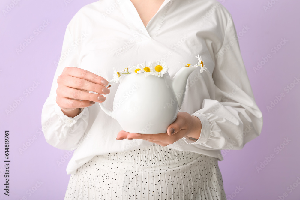 Beautiful young woman with teapot of chamomile tea and flowers near lilac wall