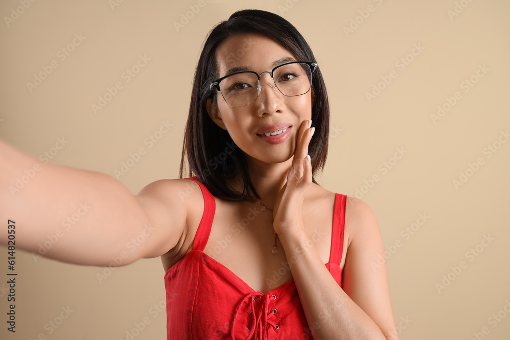 Beautiful Asian woman in stylish eyeglasses taking selfie on beige background