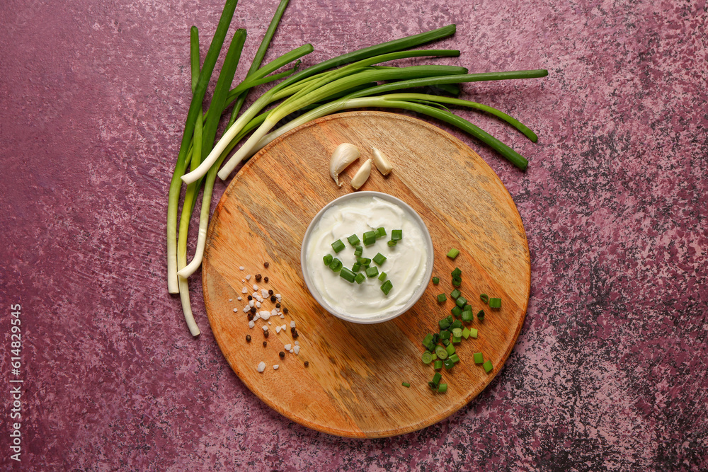 Board with bowl of tasty sour cream and sliced green onion on purple background