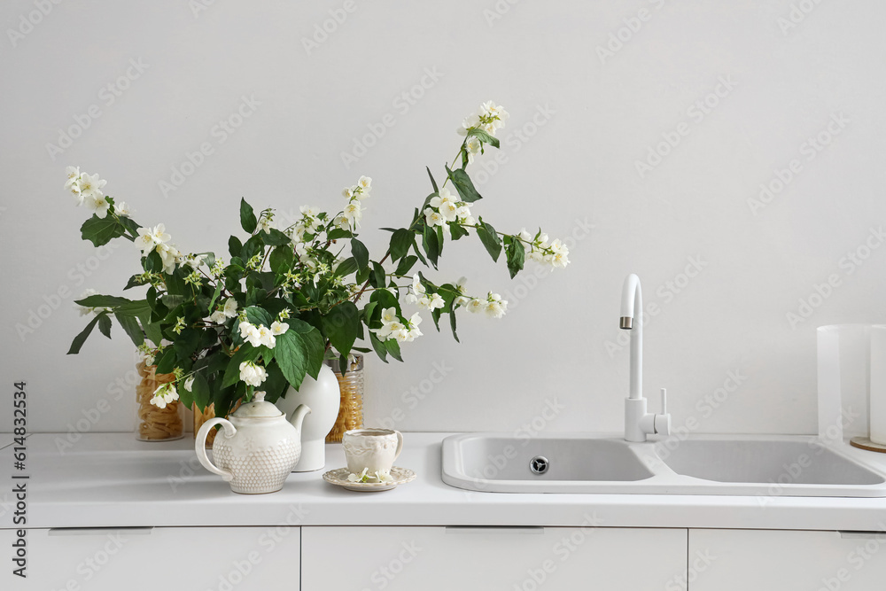 Vase with blooming jasmine flowers, teapot and cup of tea on white kitchen counter