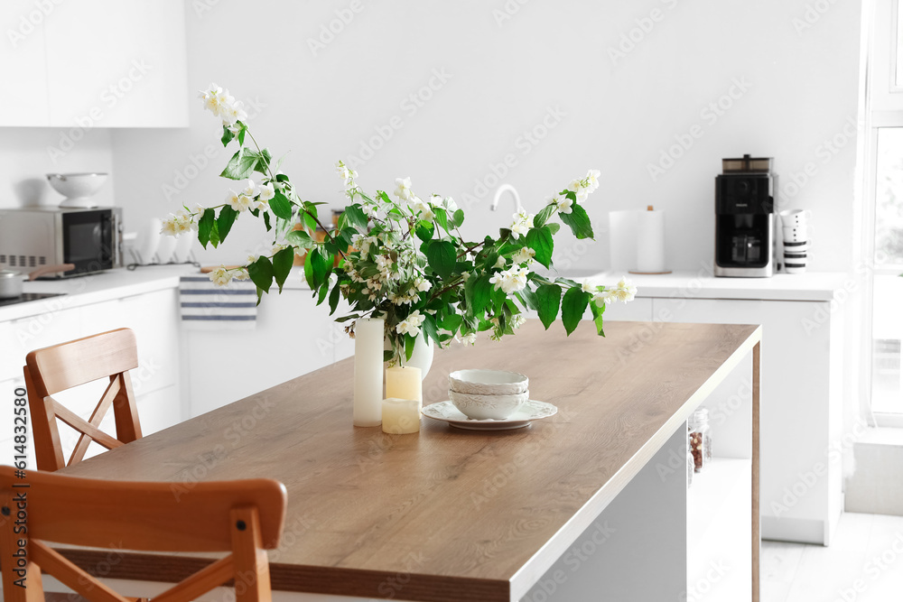 Vase with blooming jasmine flowers, candles and bowls on wooden table in modern kitchen