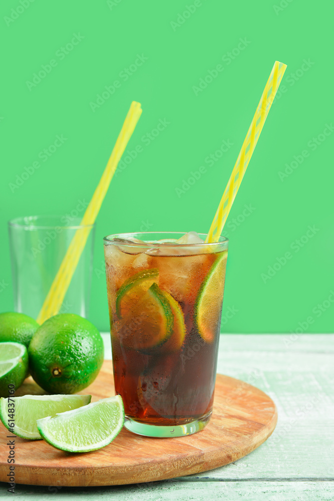 Board with glass of cold Cuba Libre cocktail and limes on green wooden table