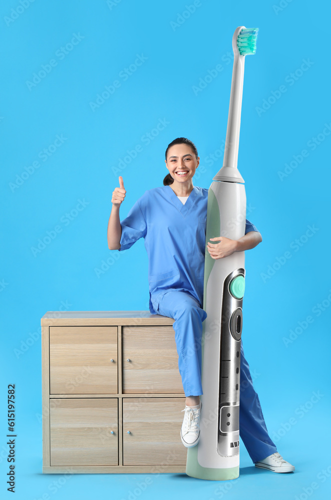 Female dentist with big electric toothbrush showing thumb-up on blue background