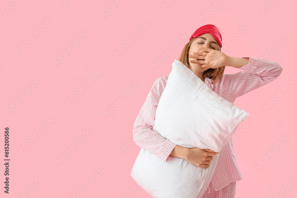 Young woman in pajamas with pillow yawning on pink background