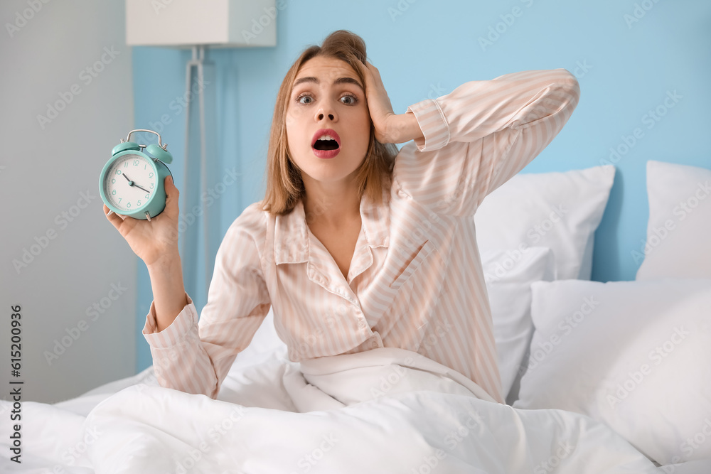 Shocked young woman with alarm clock sitting on bed in morning