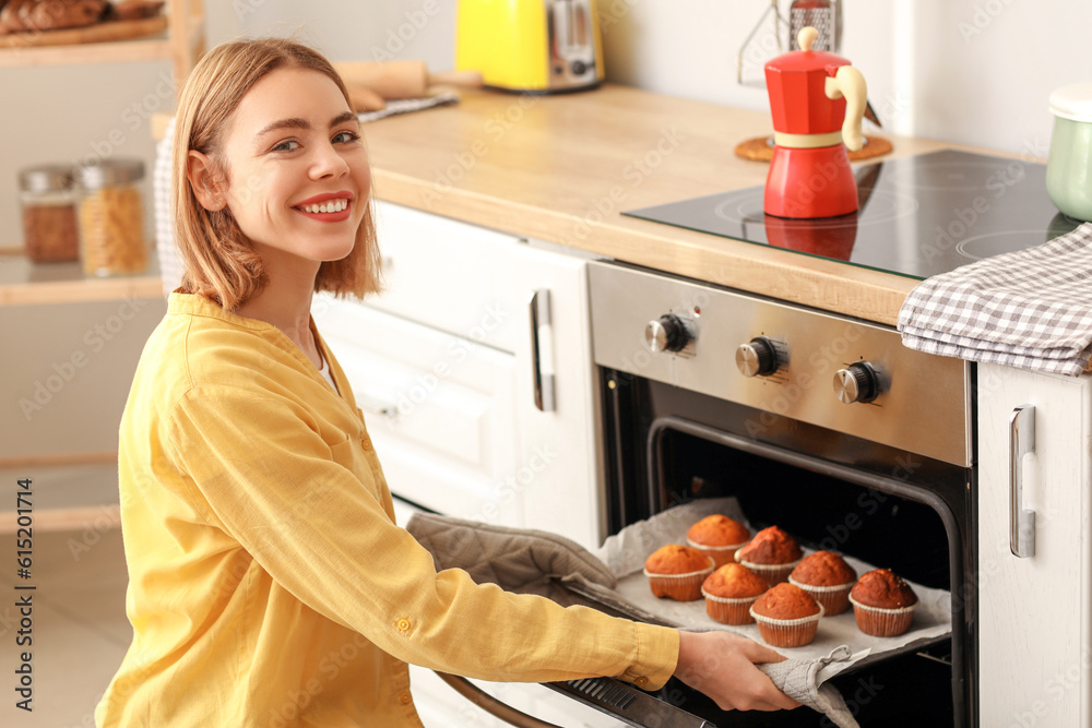 Young woman taking tray with cupcakes from oven in kitchen