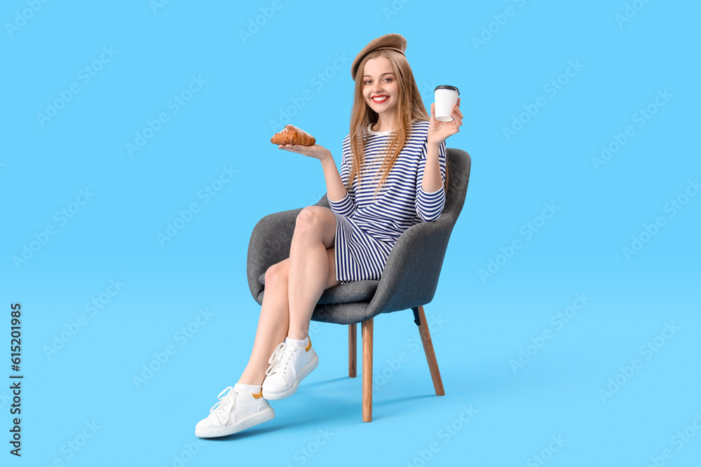 Young woman with croissant and cup of coffee sitting in armchair on blue background