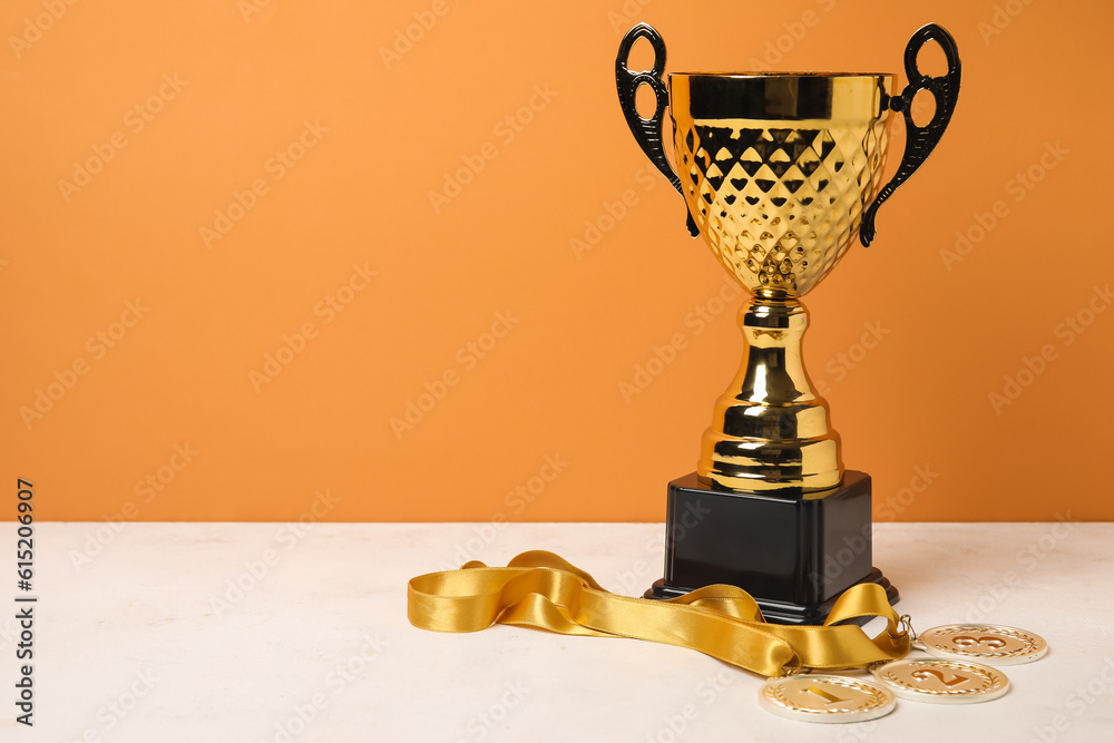 Gold cup with prize medals on table near orange wall