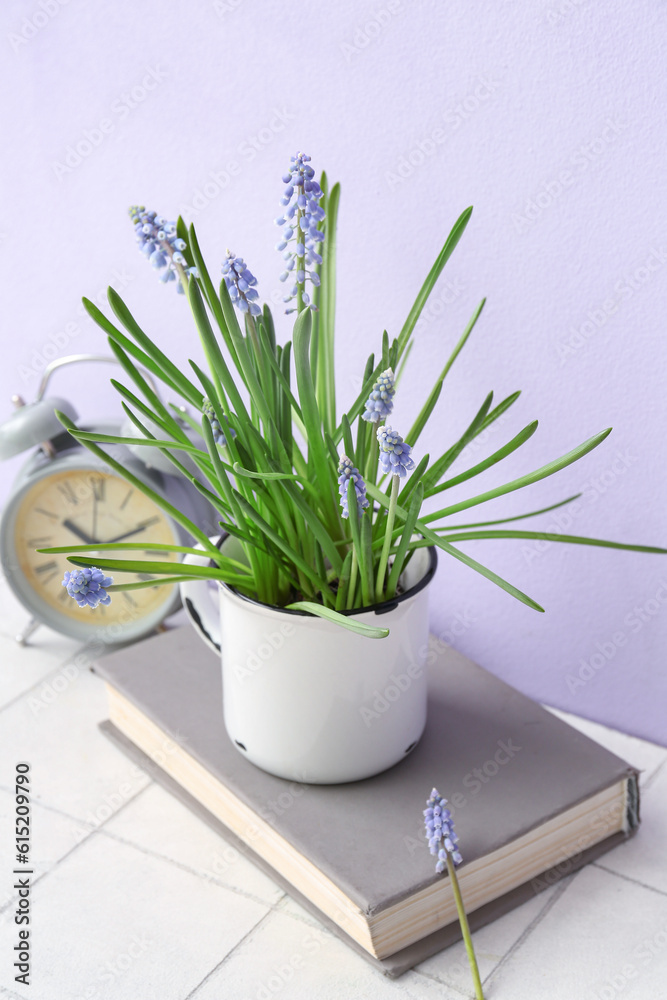 Cup with beautiful Muscari flowers, alarm clock and book on tiled table near lilac wall