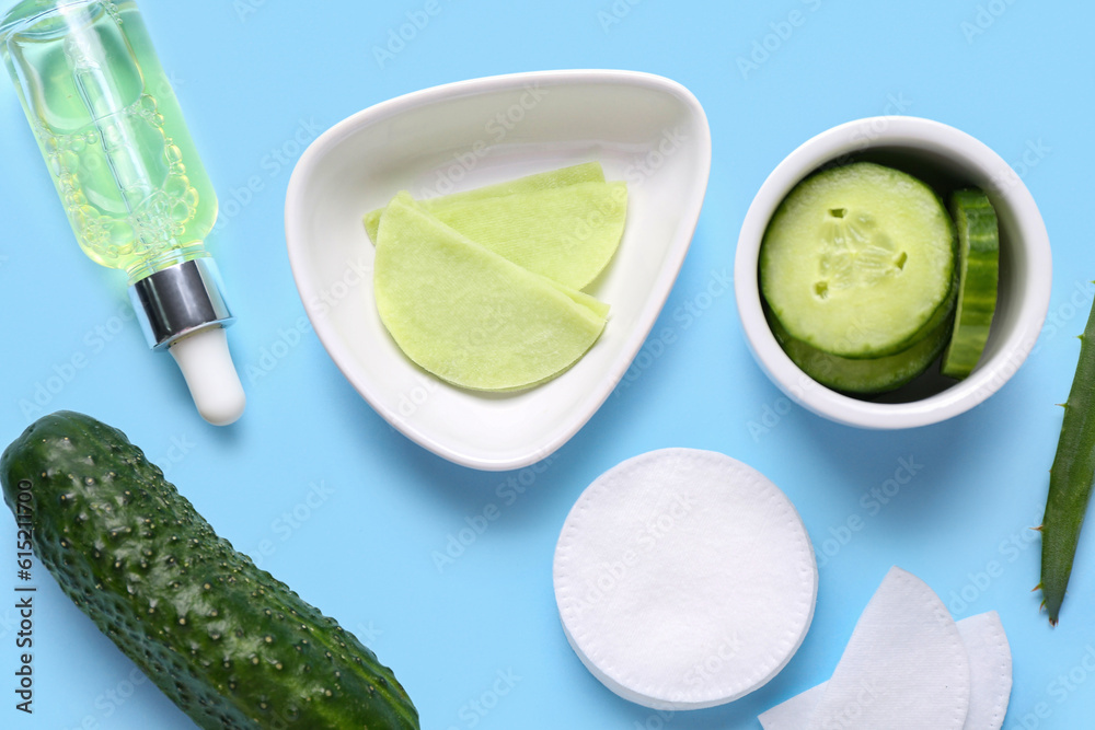 Bowls with cotton under-eye patches, cucumber and dropper bottle on blue background