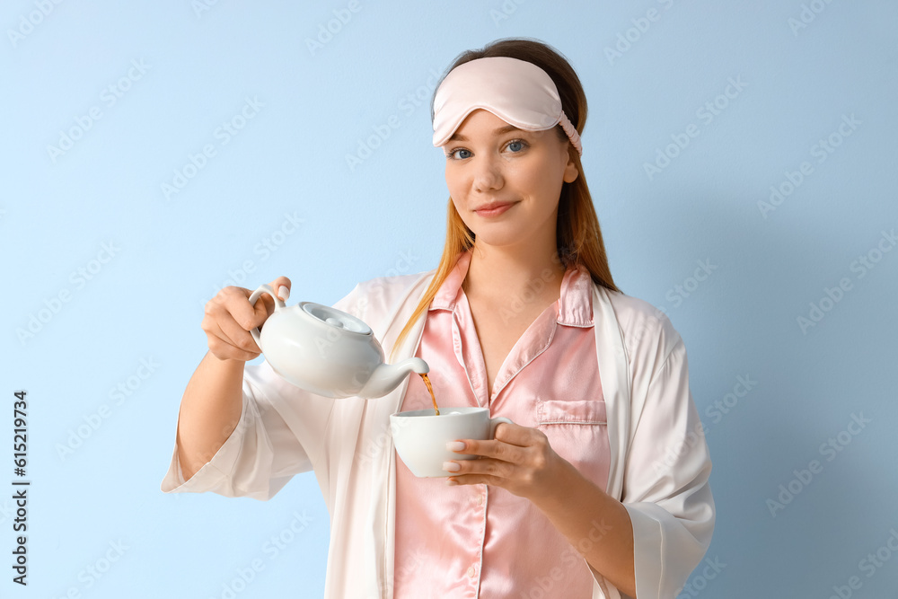 Young woman in pajamas pouring tea into cup on blue background