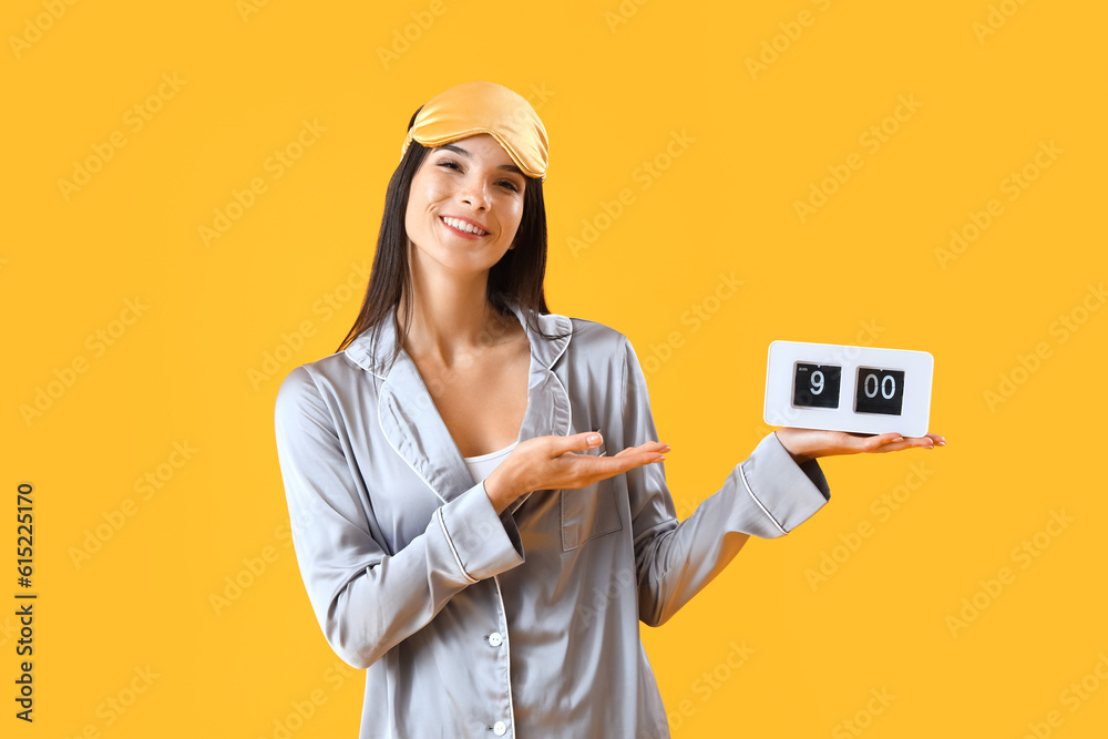 Young woman in pajamas with alarm clock on yellow background