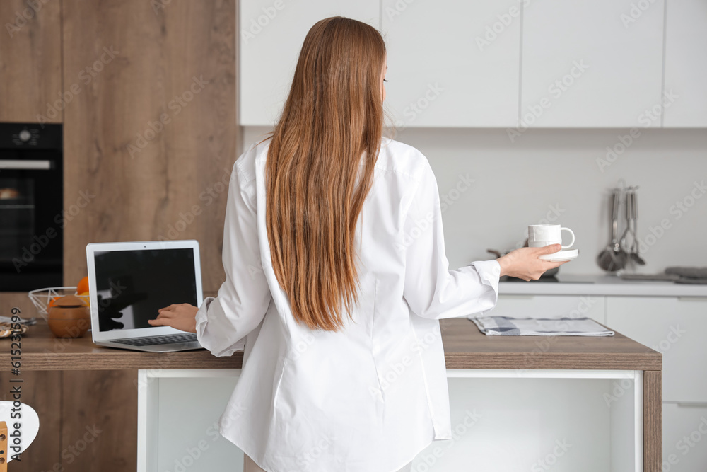 Woman working on modern laptop and holding cup of coffee in light kitchen