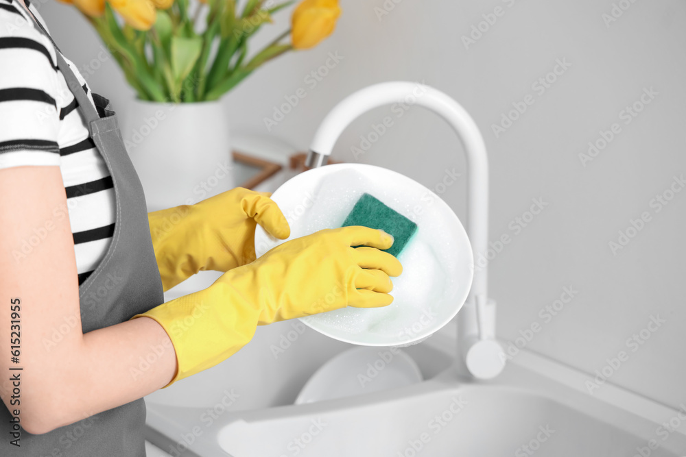 Woman in yellow rubber gloves washing plate with sponge in light kitchen