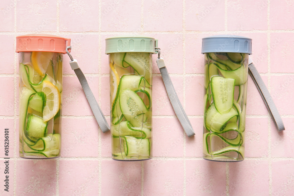 Sports bottles of infused water with cucumber slices on pink tile background