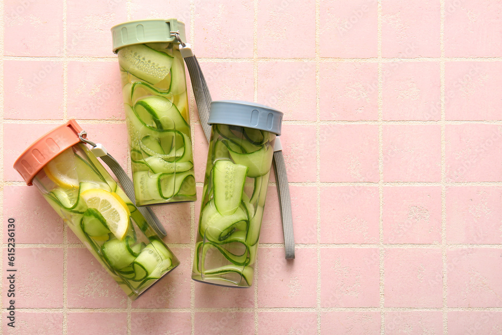 Sports bottles of infused water with cucumber slices on pink tile background