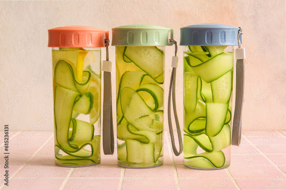 Sports bottles of infused water with cucumber slices on pink tile table