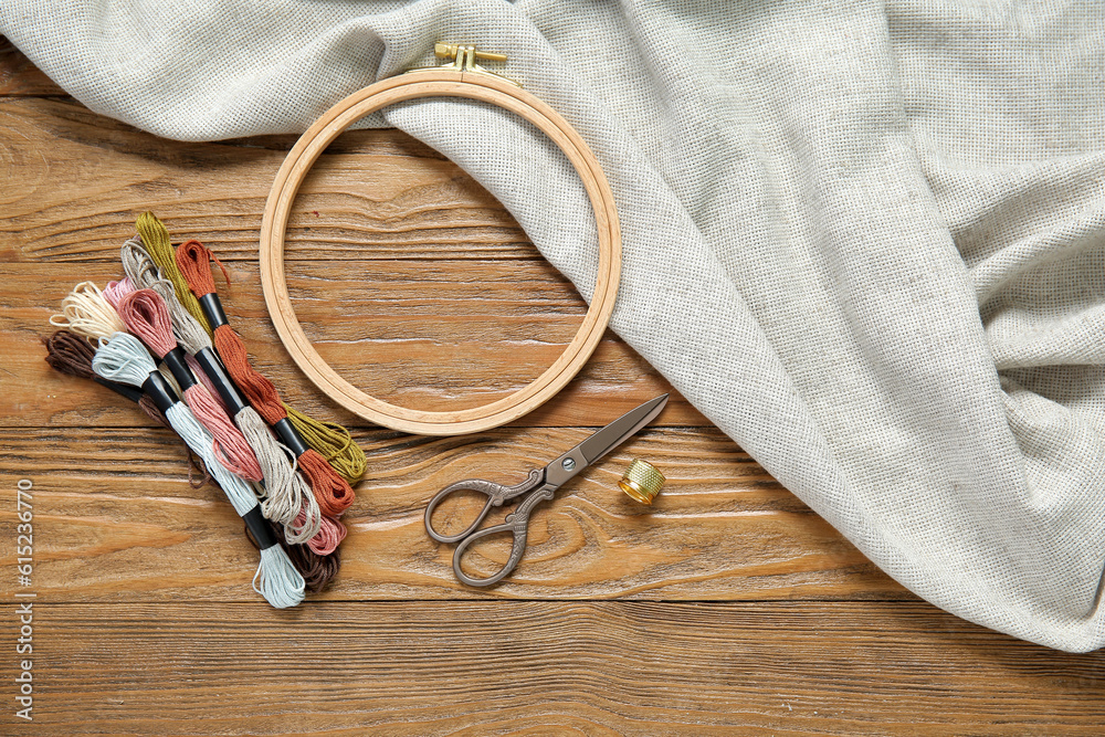 Embroidery hoop with mouline threads, scissors, thimble and canvas on brown wooden background