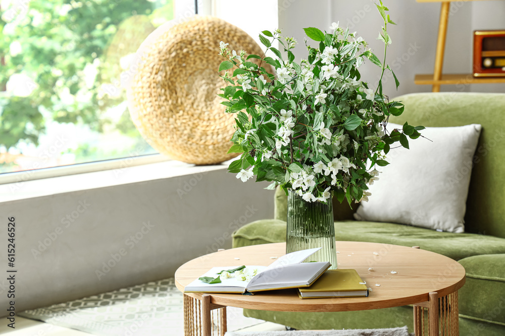 Vase with blooming jasmine flowers and books on wooden coffee table in living room