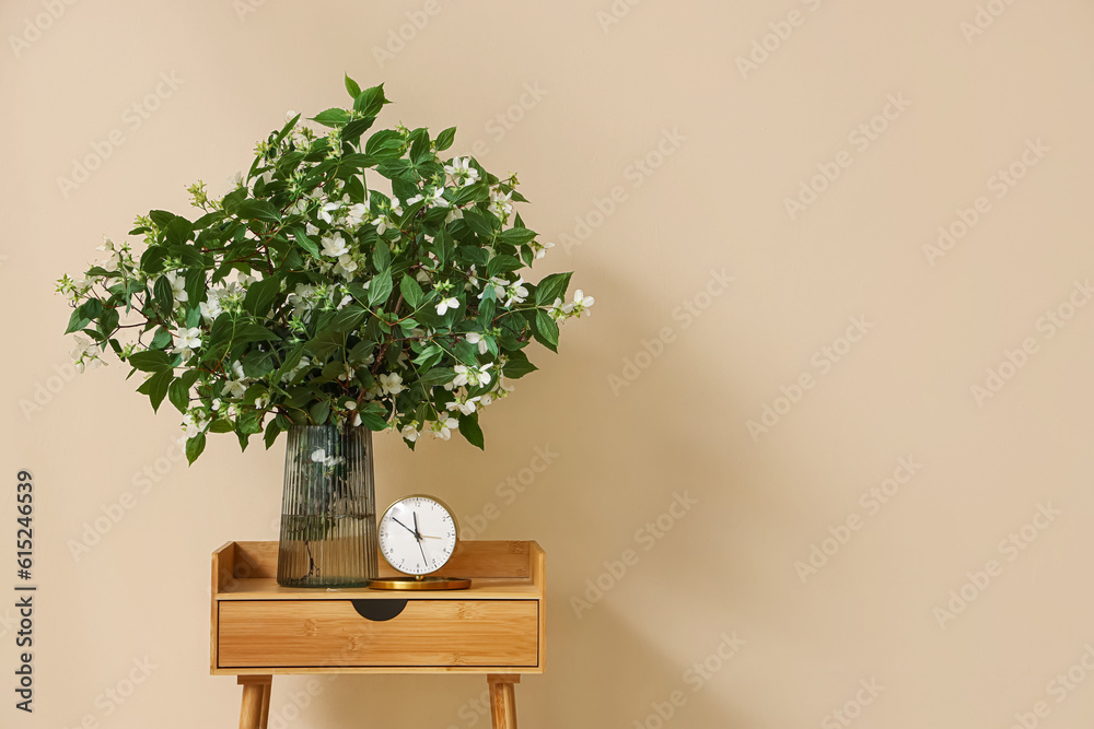 Vase with blooming jasmine flowers and alarm clock on wooden table near beige wall