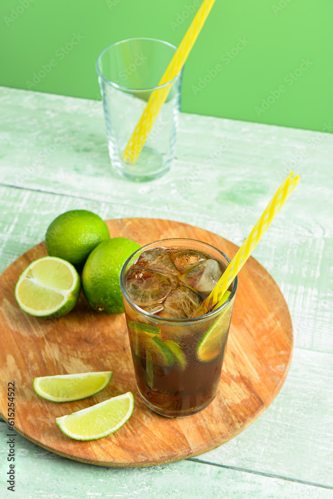 Board with glass of cold Cuba Libre cocktail and limes on green wooden table