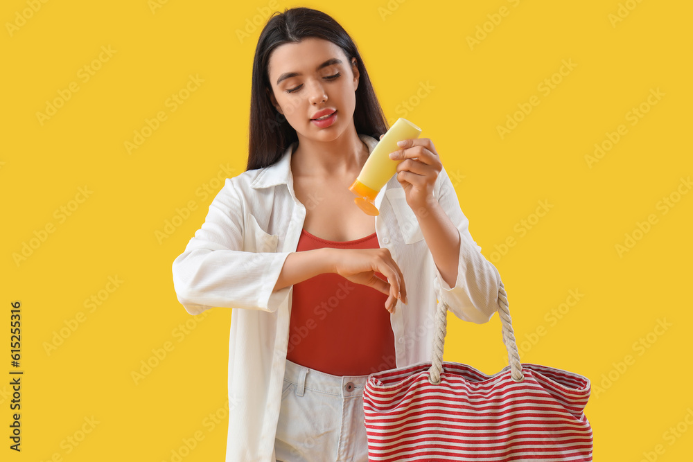 Young woman with beach bag applying sunscreen cream on yellow background