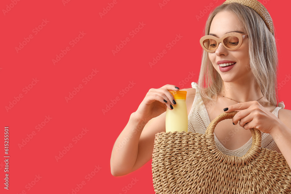 Young woman putting sunscreen cream in bag on red background