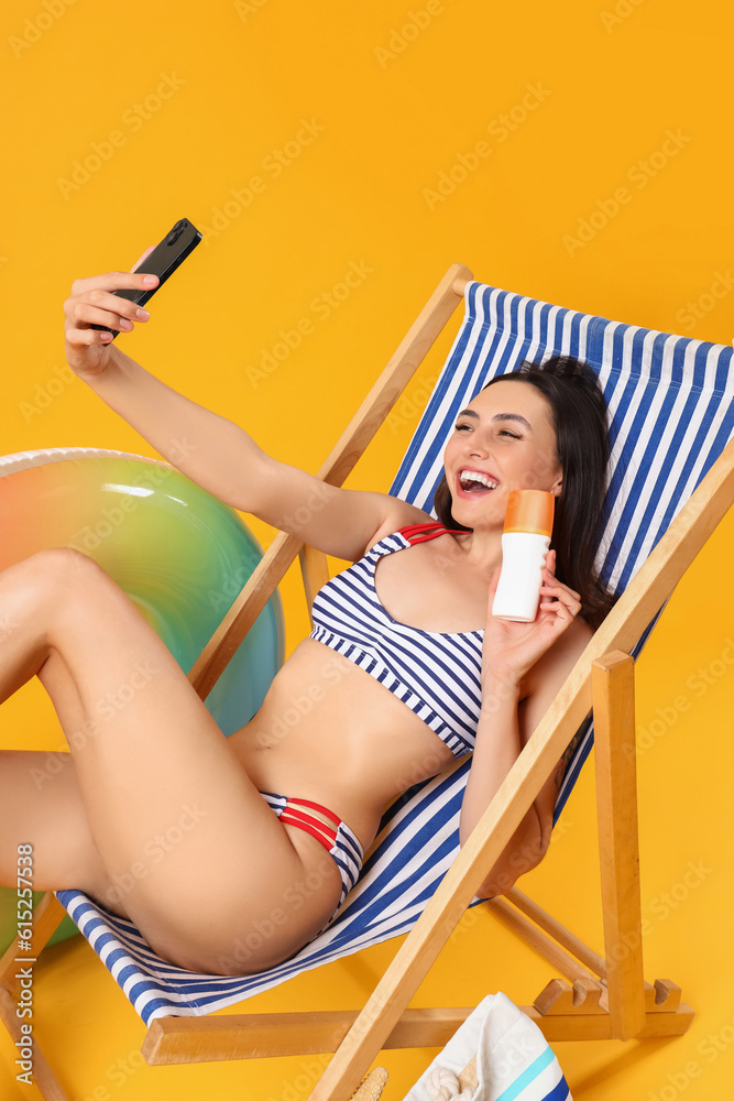 Young woman with sunscreen cream taking selfie in deck chair on yellow background