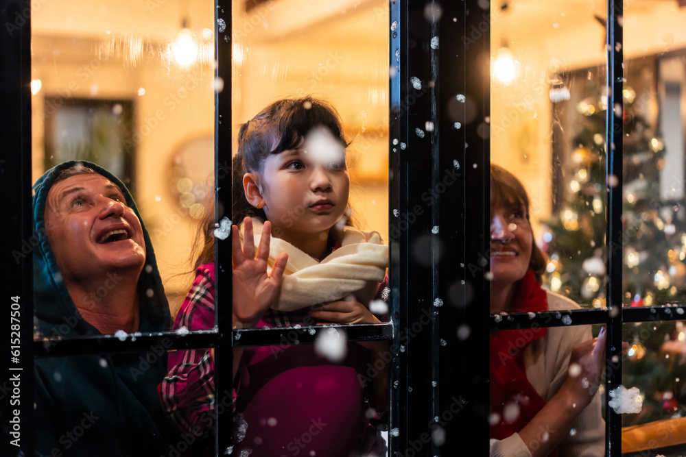 Adorable child looking at the window and first snow flakes with family. 