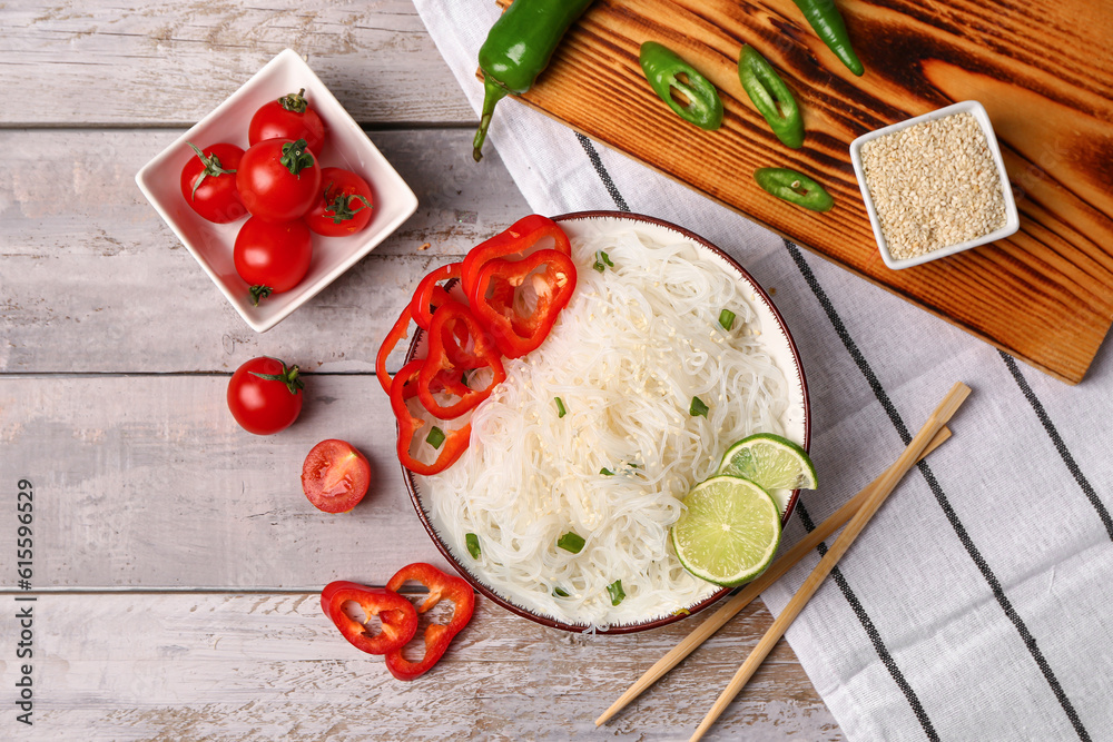 Composition with bowl of tasty rice noodles, vegetables and sesame seeds on light wooden background