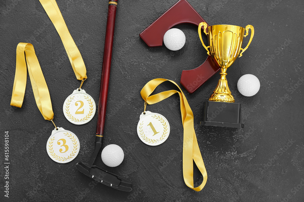 Gold cup with prize medals, golf club and balls on dark background