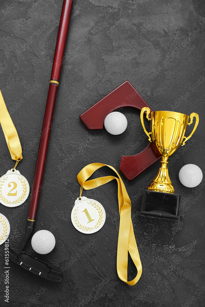 Gold cup with prize medals, golf club and balls on dark background