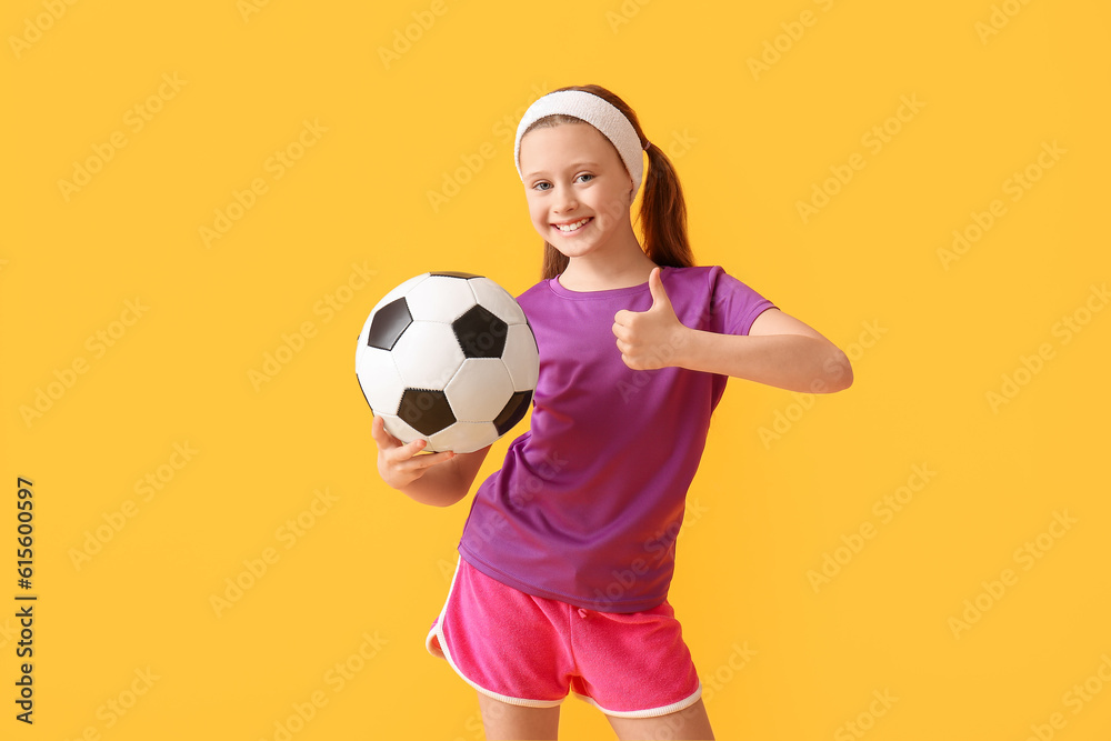 Sporty little girl with soccer ball showing thumb-up on yellow background