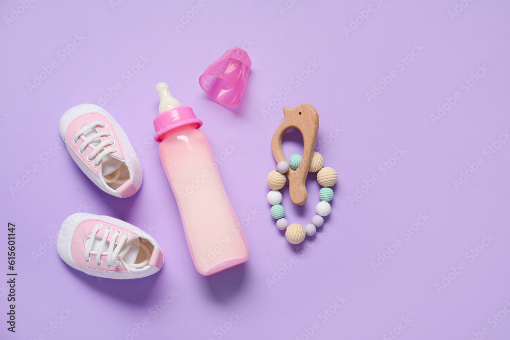 Bottle of milk for baby with rattle and booties on lilac background