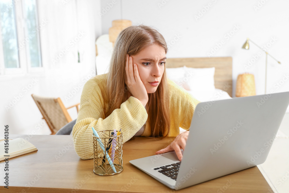 Thoughtful young woman with laptop at table in bedroom