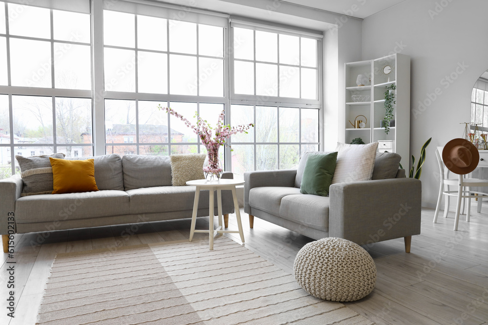 Interior of living room with grey sofas and blooming sakura branches on coffee table near big window