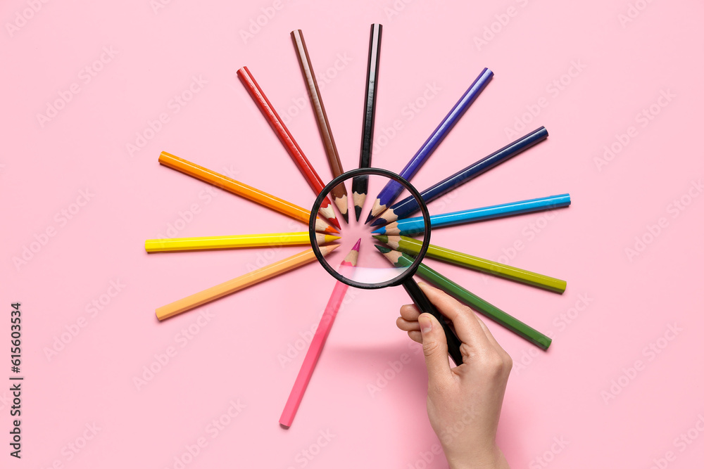 Female hand with magnifier and frame made of colorful pencils on pink background