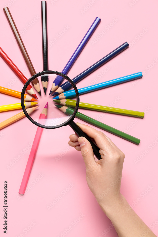 Female hand with magnifier and frame made of colorful pencils on pink background