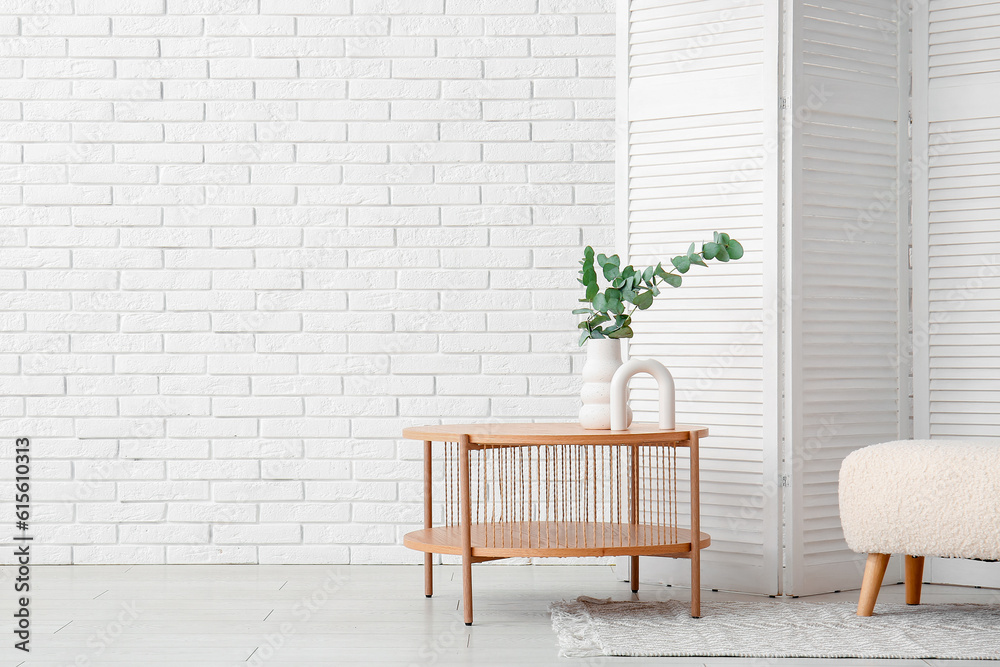 Wooden coffee table with eucalyptus branches in vase, folding screen and pouf near white brick wall