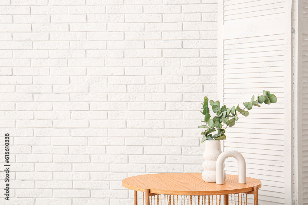 Wooden coffee table with eucalyptus branches in vase and folding screen near white brick wall