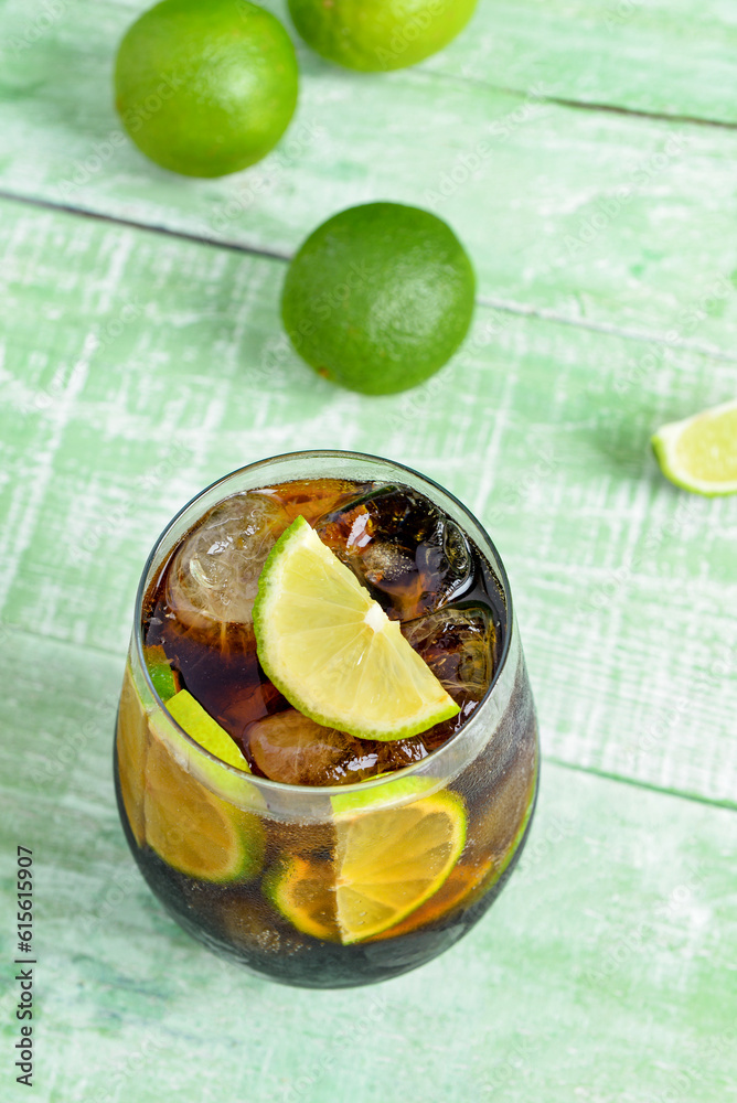 Glass of cold Cuba Libre cocktail and limes on green wooden table